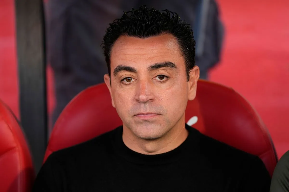 Close-up of a man with dark hair in a black shirt sitting on a red stadium bench, looking thoughtful.