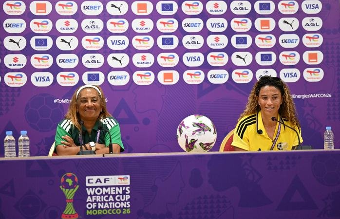 Two female footballers sit at a press conference desk with a ball between them, purple backdrop bearing sponsor logos for the CAF Women's Africa Cup of Nations Morocco 2026.