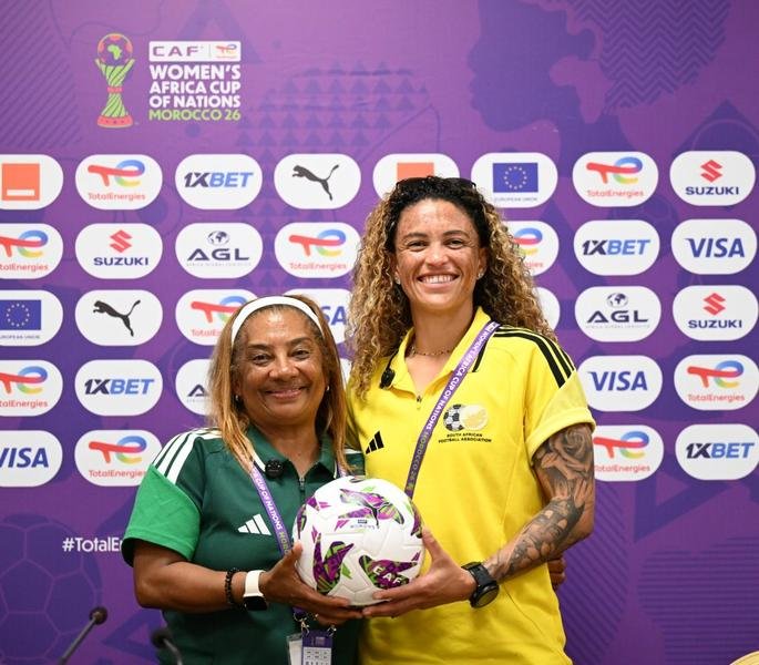 Two women pose with a soccer ball at a CAF Women's Africa Cup of Nations event; one in green, one in yellow with medals.