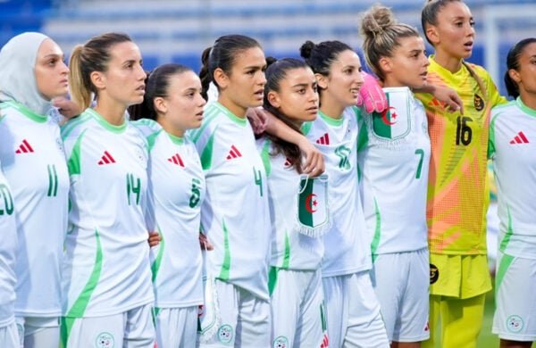 Women’s soccer players in white and green uniforms stand in a line with arms around each other’s shoulders, ready for a team photo or moment of unity; one player wearing a light blue hijab is visible on the left and a yellow-clad goalkeeper is toward the right.