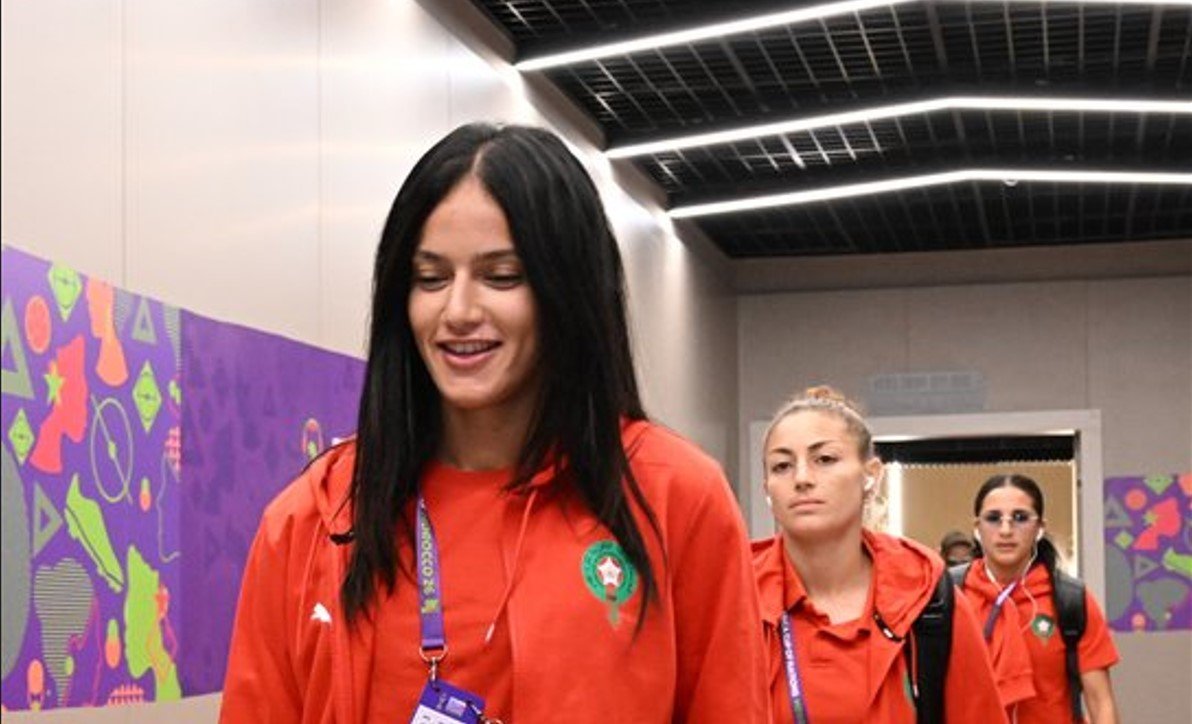 A woman with long dark hair wearing a red shirt and lanyard walking through a colorful hallway, smiling slightly.