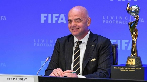 FIFA president seated at a desk during a press conference, smiling, wearing a dark suit and striped tie, with a microphone and a trophy beside him on the right.
