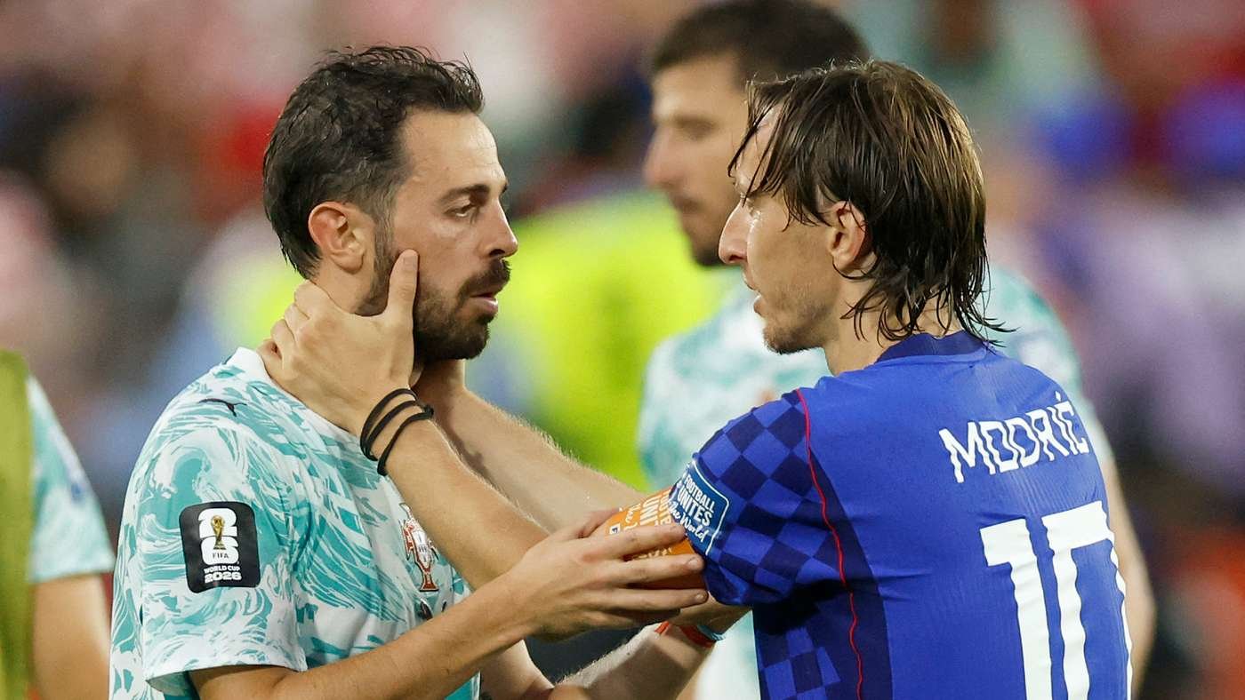 Two male soccer players on the field in a close conversation; one in a light-teal patterned jersey, the other in a blue jersey with number 10 on back.