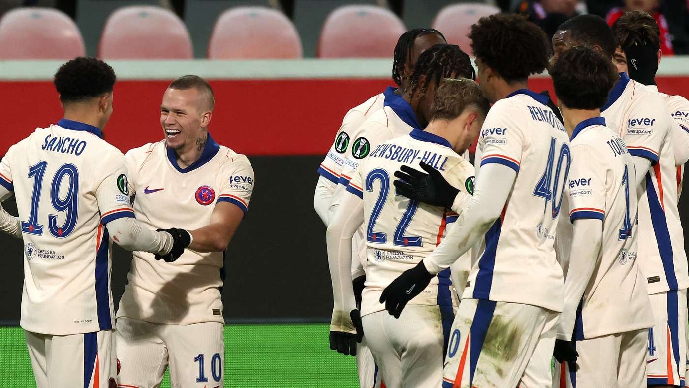 Soccer players in white uniforms celebrate a goal, hugging and smiling on the pitch.