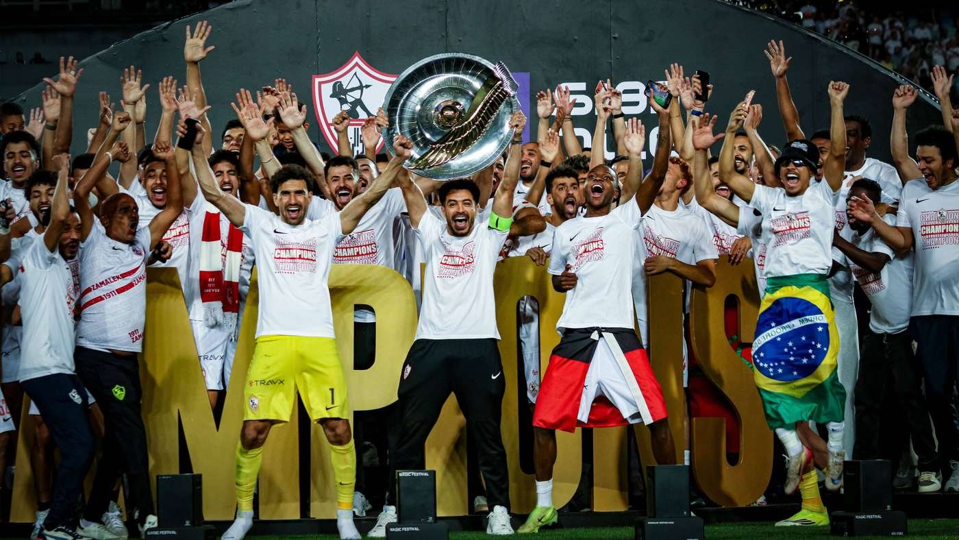 A team of soccer players celebrates on stage, lifting a large trophy as fans cheer behind them, with a Brazil flag visible on one player's shorts.