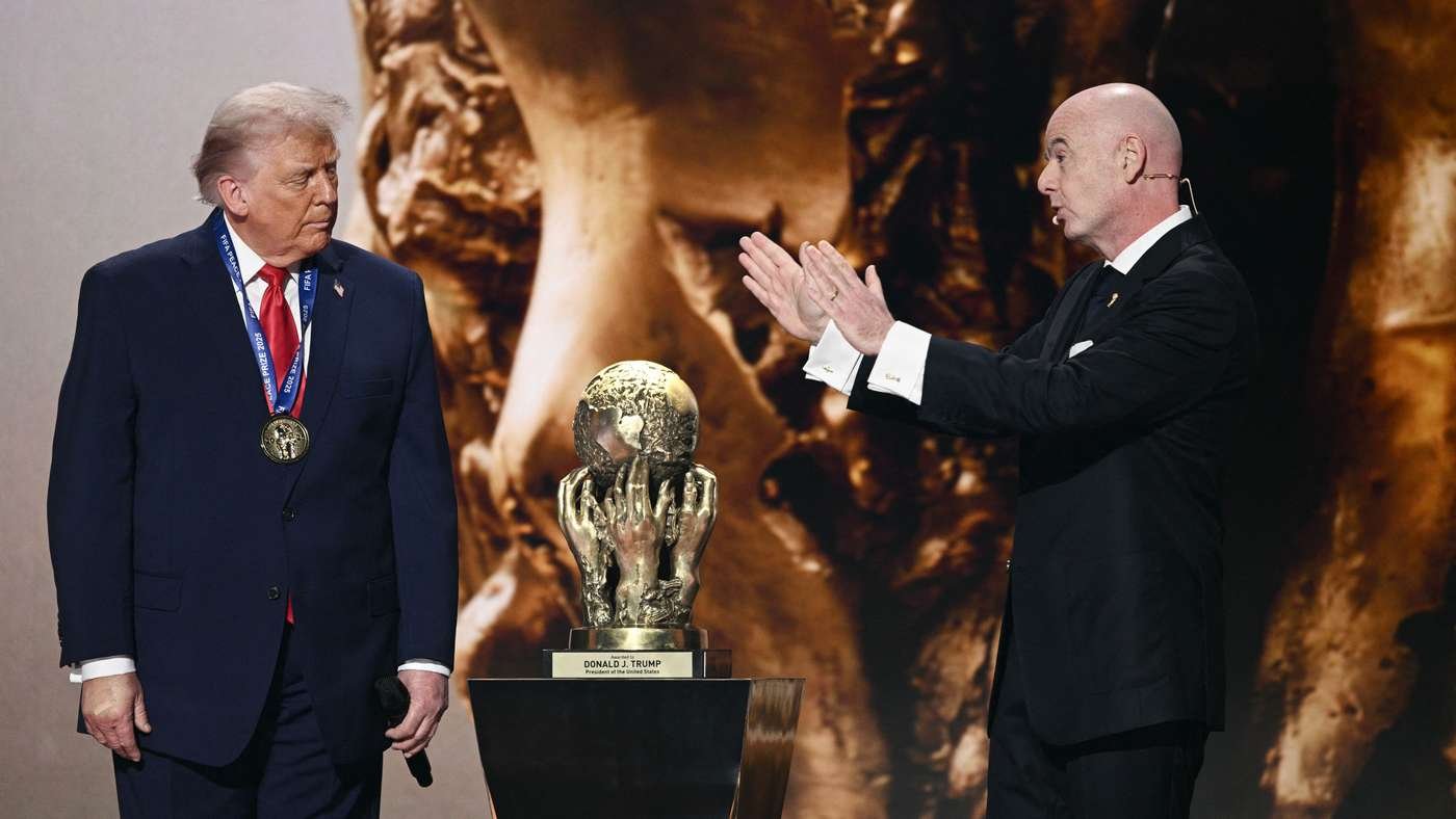 Two men in formal suits on stage with a gold globe trophy on a pedestal between them, one wearing a medal.
