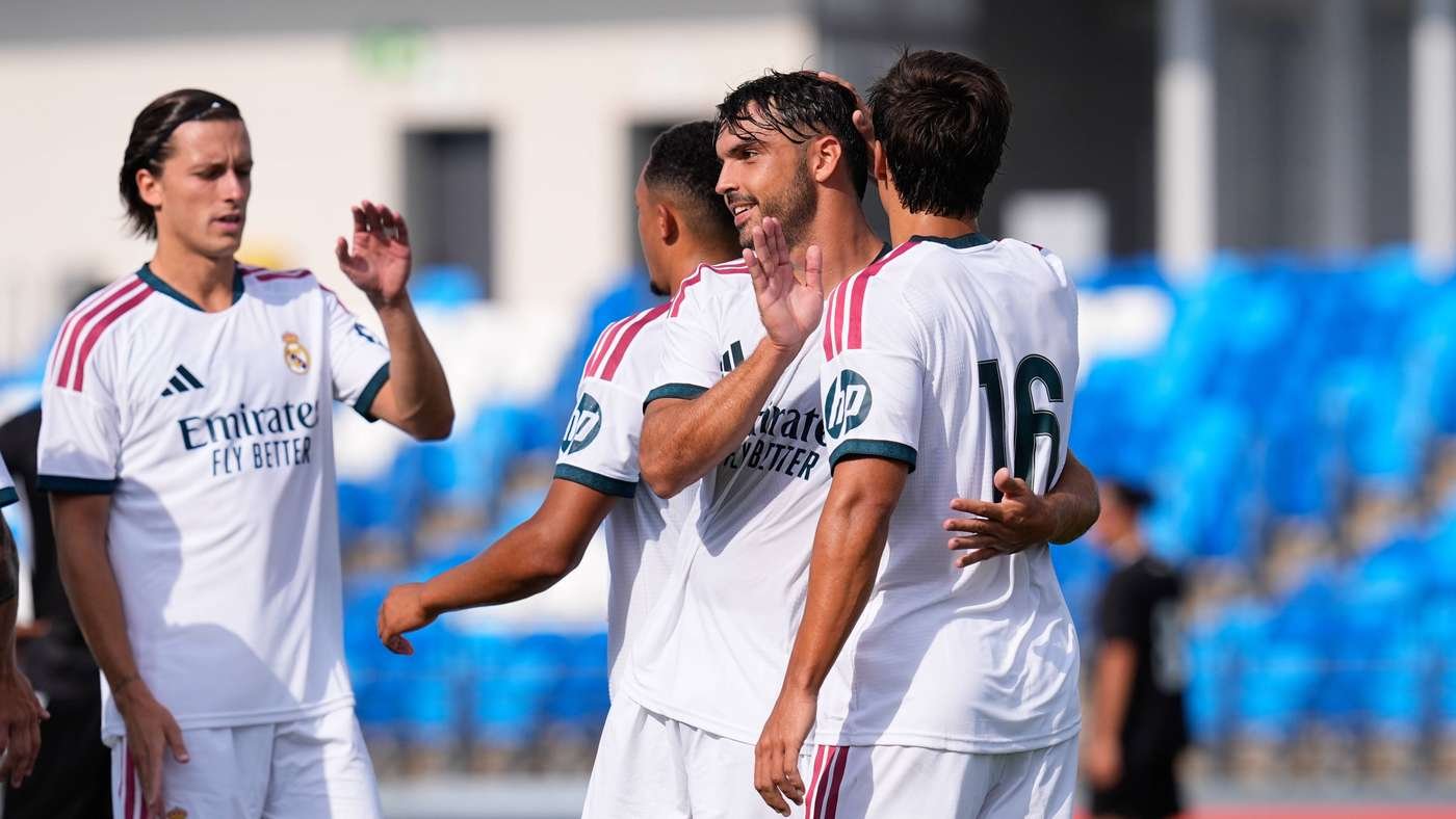 Real Madrid players celebrate on the soccer field, embracing and high-fiving in white kits with pink accents, blue seats in the background.