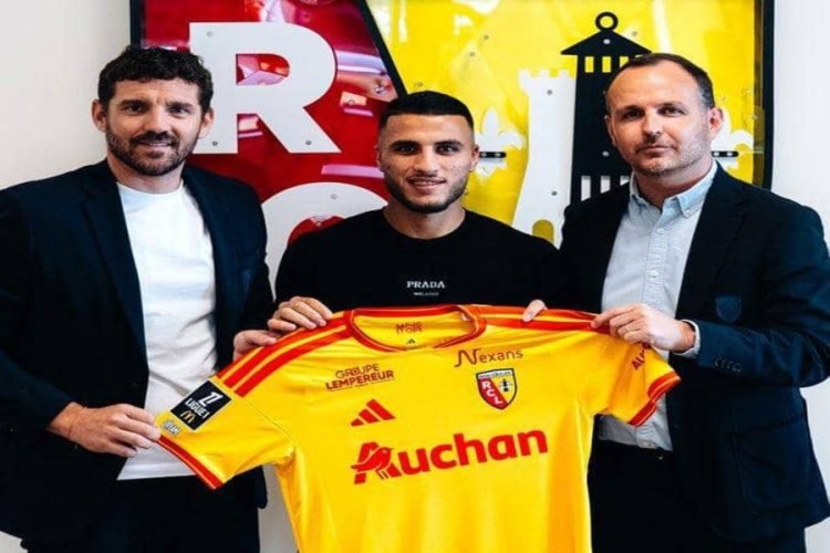 Three men in suits and casual wear pose together, holding a yellow football jersey with red sponsor logos (Auchan) and club crest, in front of a colorful wall art.