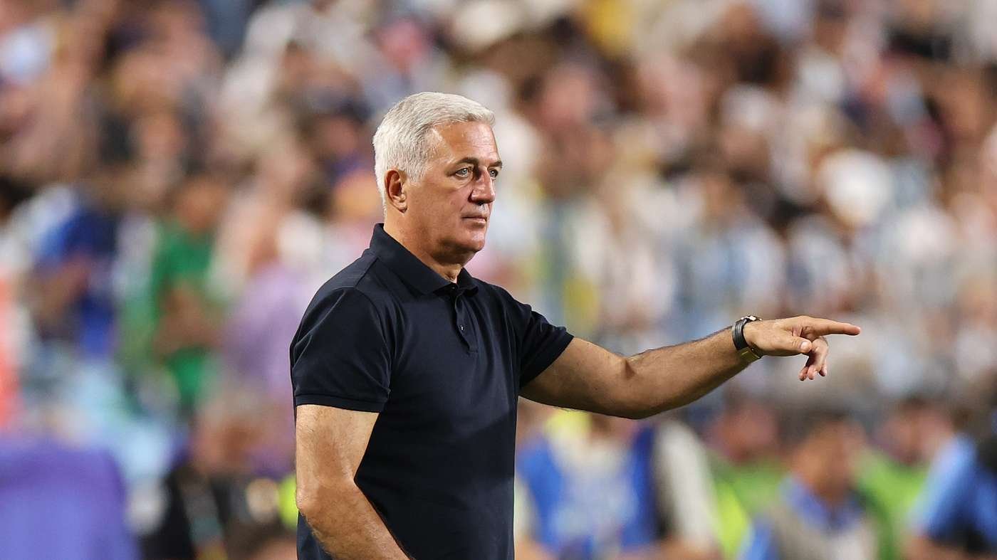 Sports coach in a navy polo points to the right on the sideline with a blurred crowd in the stands behind him.