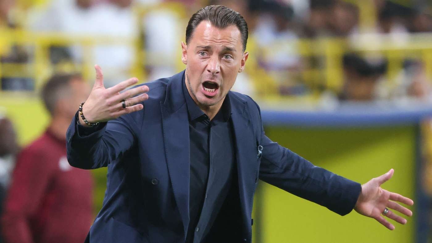 Coach in a navy suit shouting with arms spread wide on the sideline of a stadium.