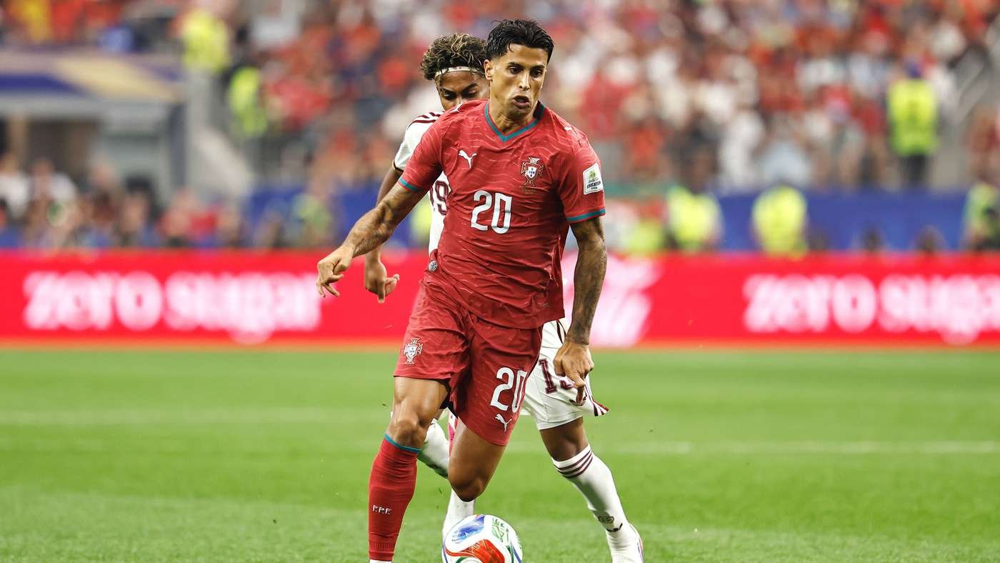 Portugal footballer wearing #20 in red kit controls the ball on a green pitch, with a white-clad defender close behind in a packed stadium