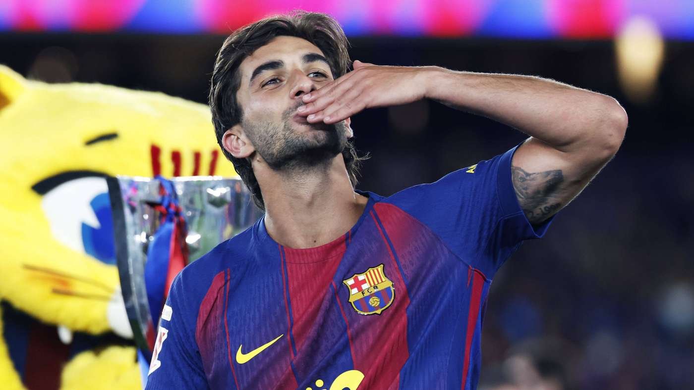 FC Barcelona player in a blue-red kit kisses his hand in celebration after lifting the trophy, with the cup visible behind him and a yellow mascot in the background.