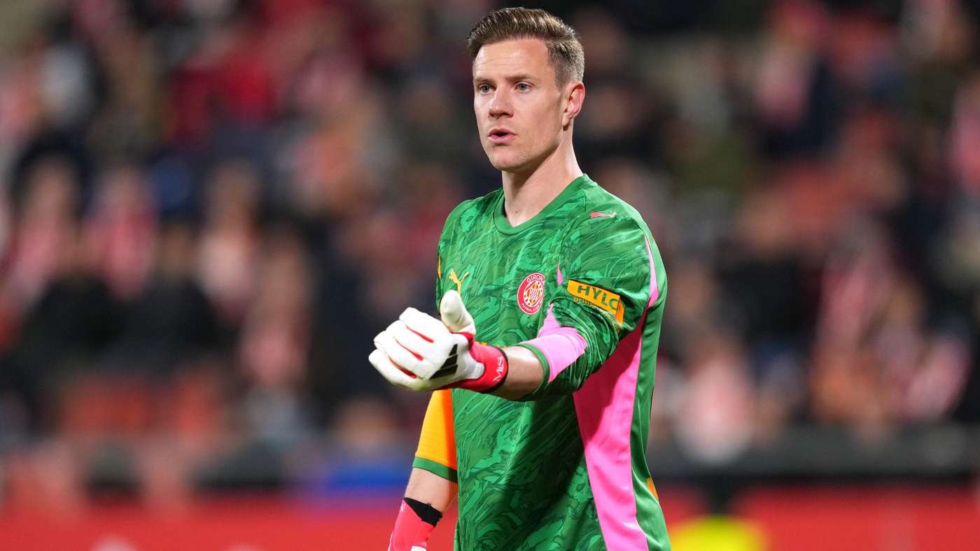 Goalkeeper in a green jersey with pink side panels and an orange sponsor patch on the sleeve, wearing gloves and extending his arm during a match with a blurred crowd in the background