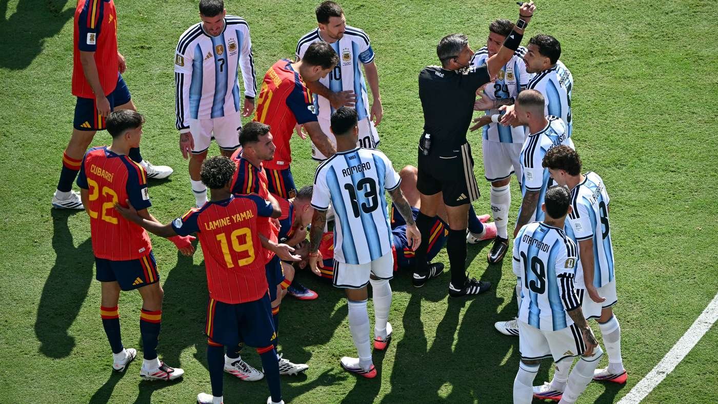 Football players from two teams gather around a referee on a green pitch, arguing as a teammate lies on the ground nearby.