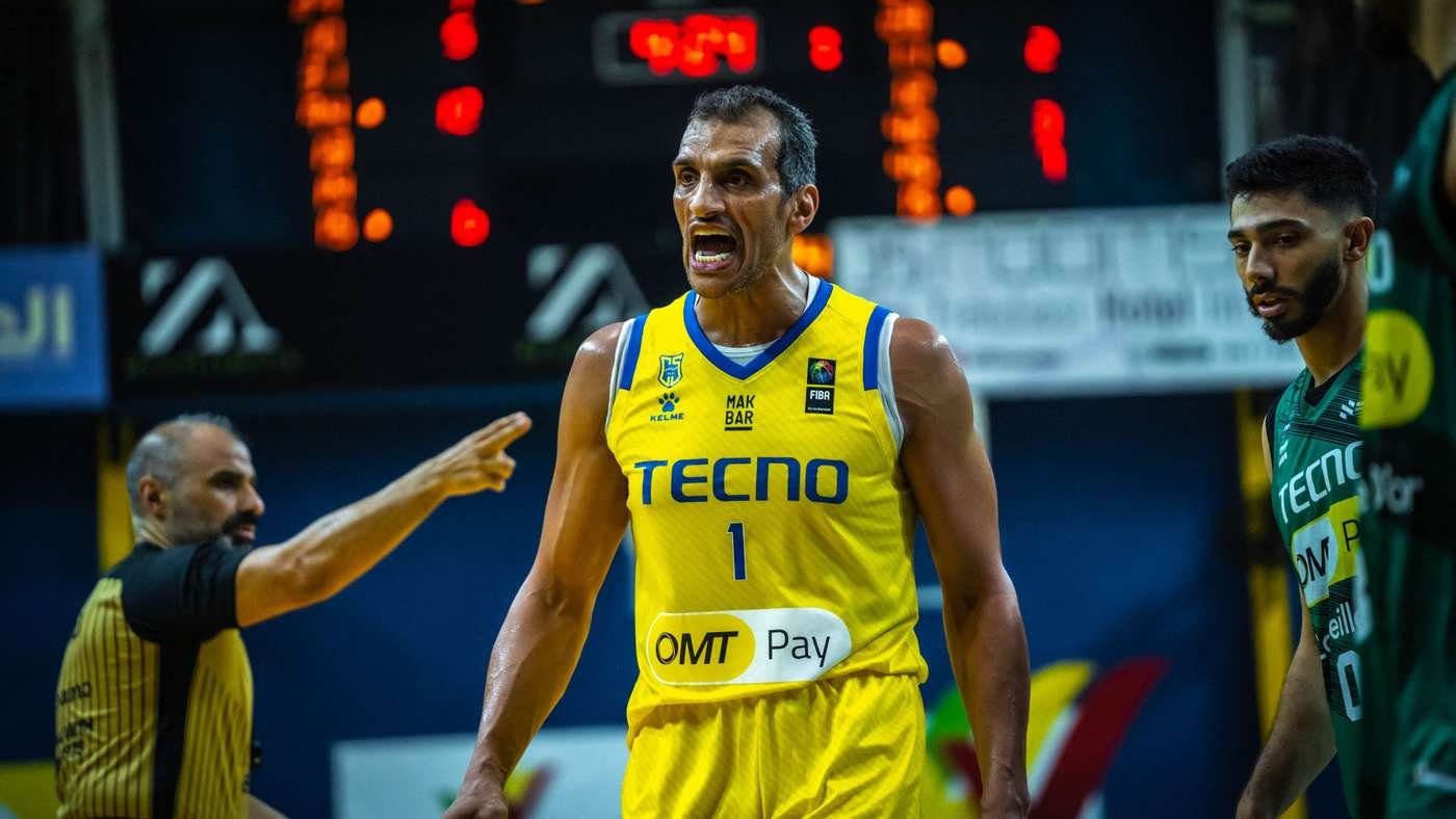 Basketball player in a yellow TECNO jersey (#1) shouting during a game, with teammates and scoreboard in the background.