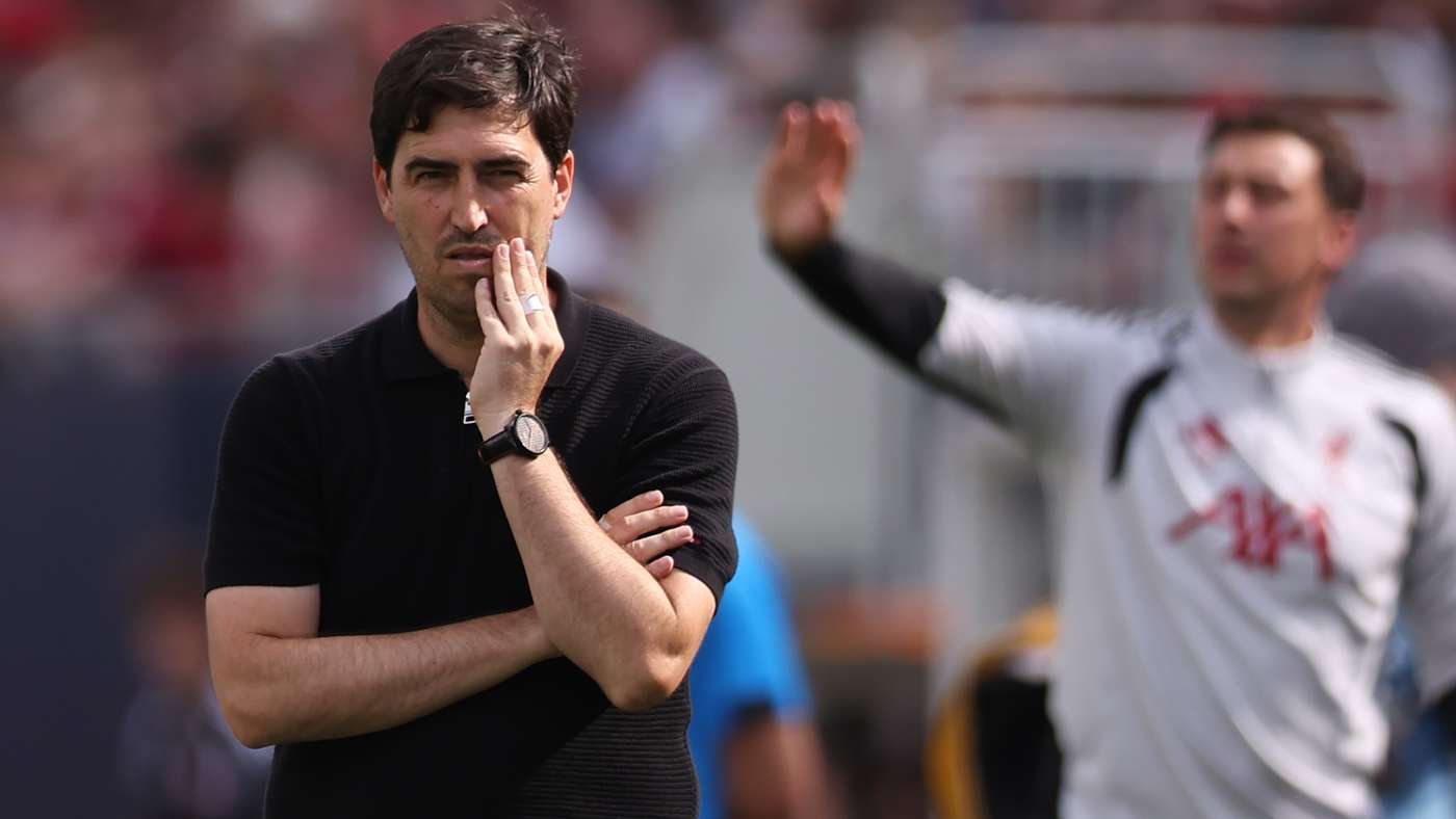 Man in a black polo rests his chin on his hand on a soccer field, looking pensive as a teammate gesticulates in the blurred background.
