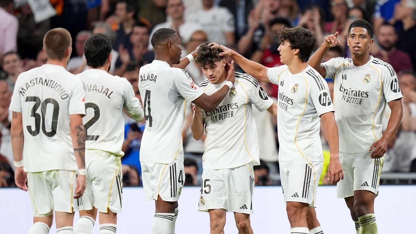 Real Madrid players in white uniforms celebrate a goal on the pitch with teammates touching heads and shoulders in a group hug.