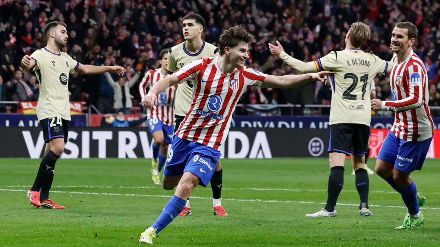 Atlético Madrid player in red-and-white stripes celebrates a goal with outstretched arms as teammates and opponents watch on a green pitch.