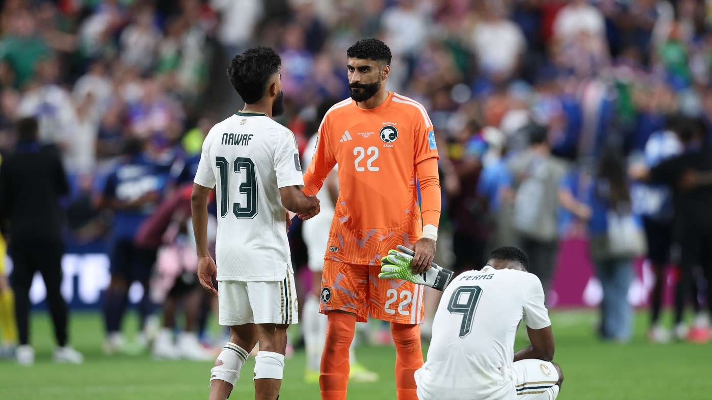 Two players in white uniforms shake hands with an orange-clad goalkeeper standing between them on a soccer field; another white-uniformed player sits on the ground nearby as a crowded stadium looms in the background.
