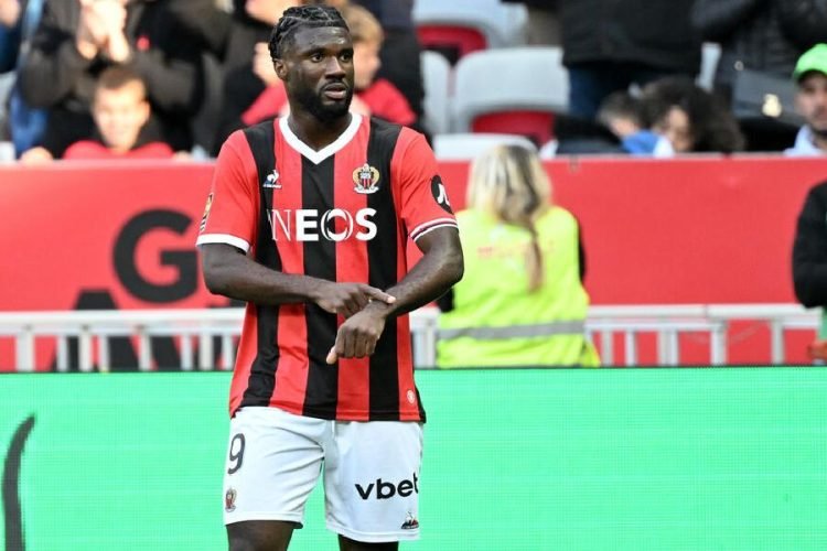 Soccer player in red-and-black striped jersey with NEOS sponsor crosses arms on the field during a match, number 9 visible on shorts.