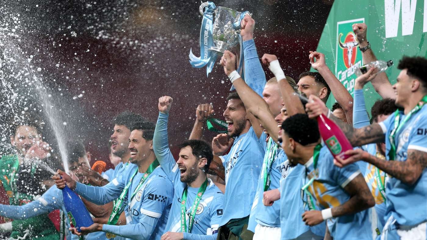 Soccer team in light-blue jerseys celebrates win, lifting a trophy as champagne sprays around them.