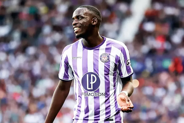 Soccer player in a purple and white striped jersey smiling on the field with a blurred crowd in the background.