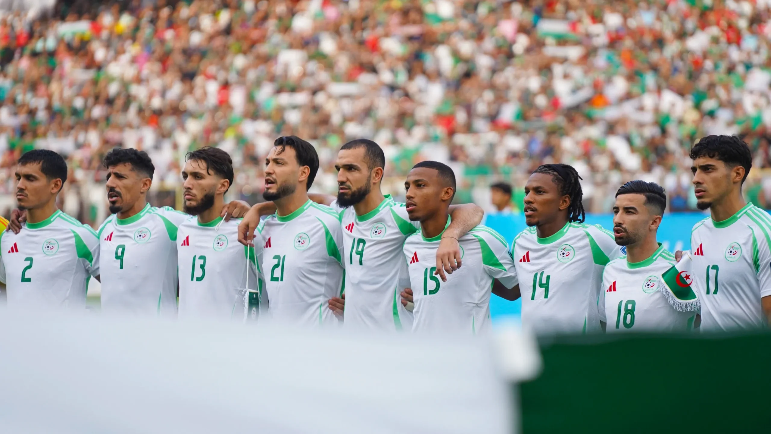 Algeria national football team lined up with arms around teammates on the pitch, crowded stadium in background.