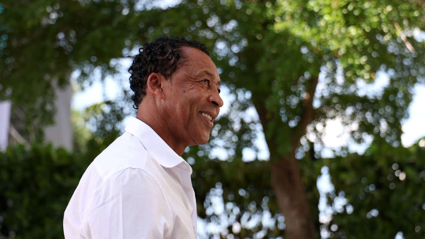 Man in a white shirt smiling in an outdoor park setting with green trees in the background, profile view.