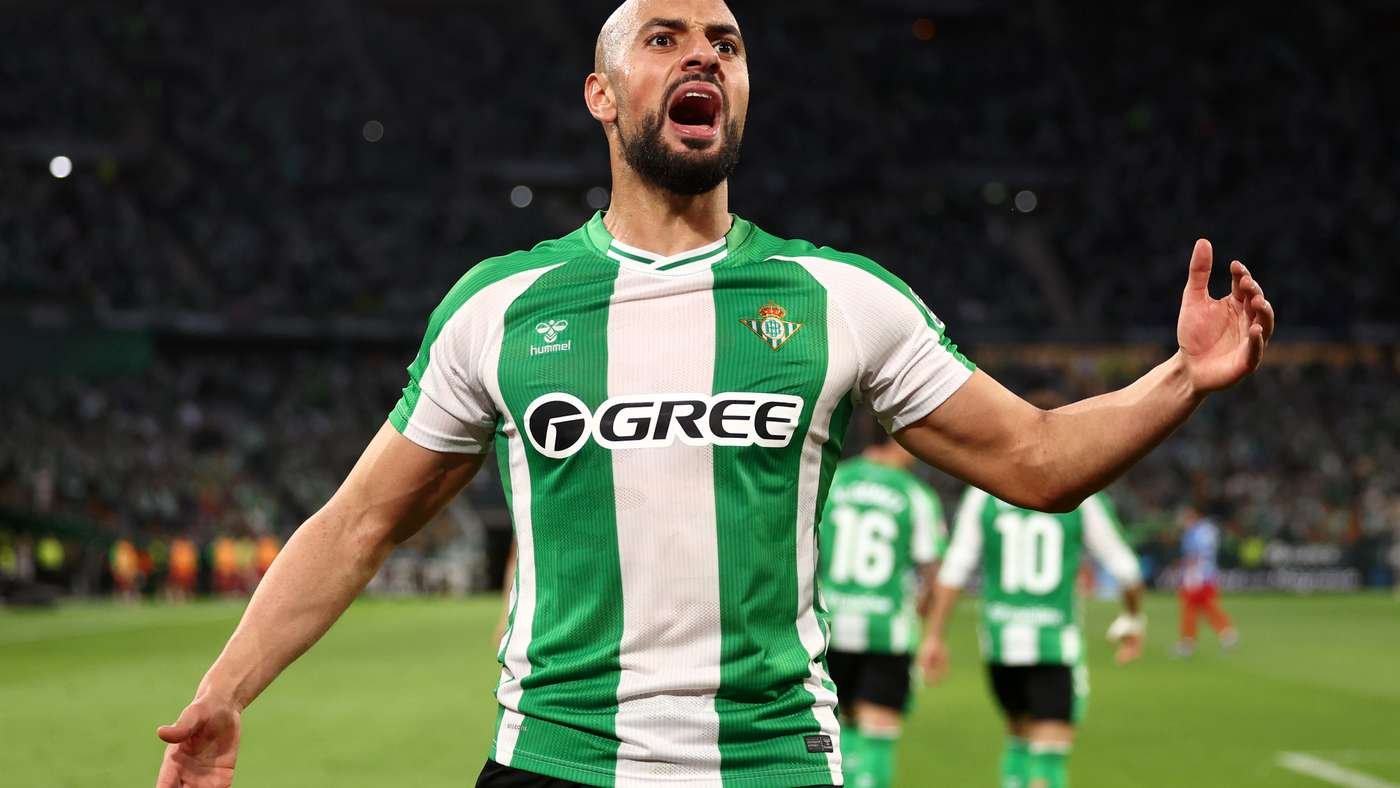Soccer player in a green and white striped jersey celebrates with arms outstretched on the pitch on a night game.