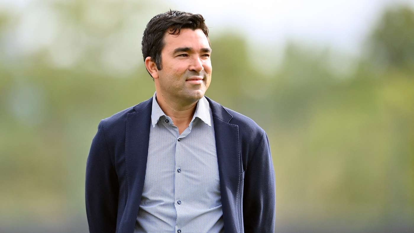 Man in a navy blazer and light checkered shirt stands outdoors with a blurred green background, looking to the right.
