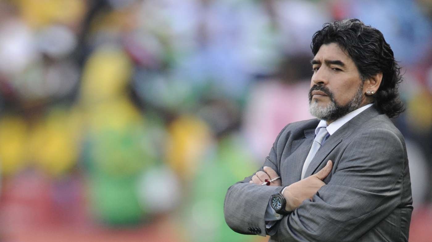 Man in a gray suit with folded arms looks contemplative outdoors, colorful blurred background behind him.