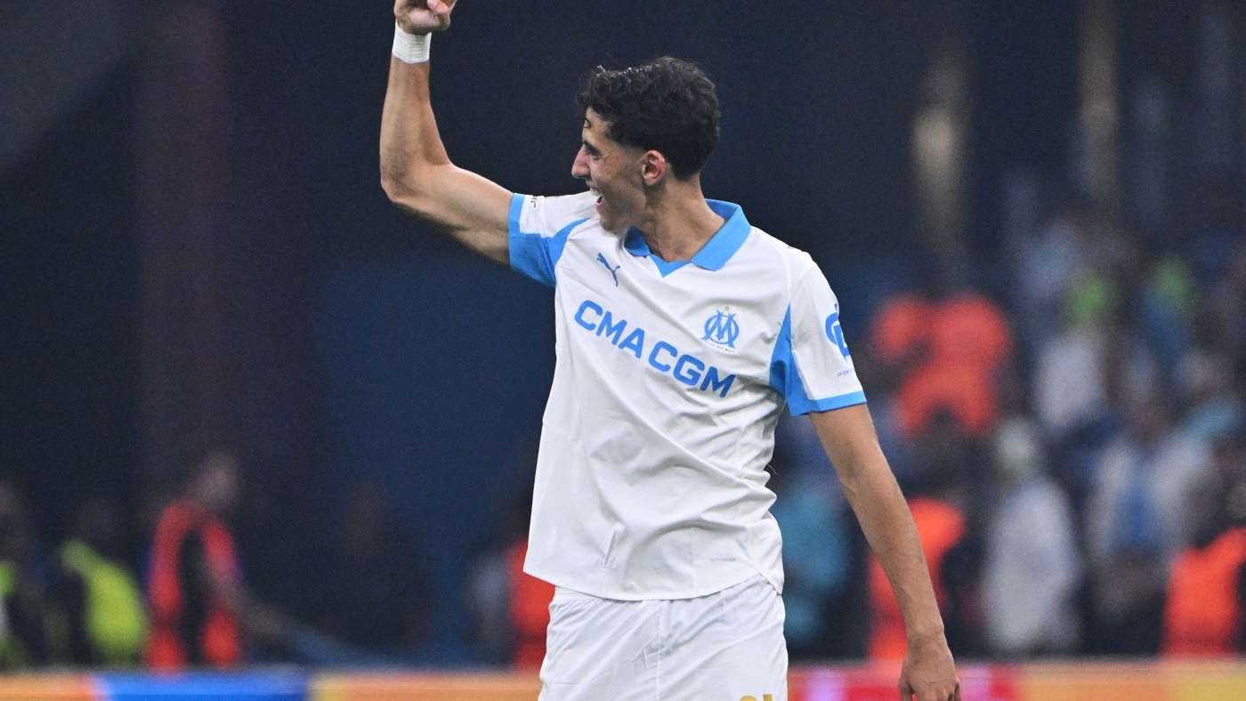Soccer player in a white and blue kit raises his right fist in celebration on the field.