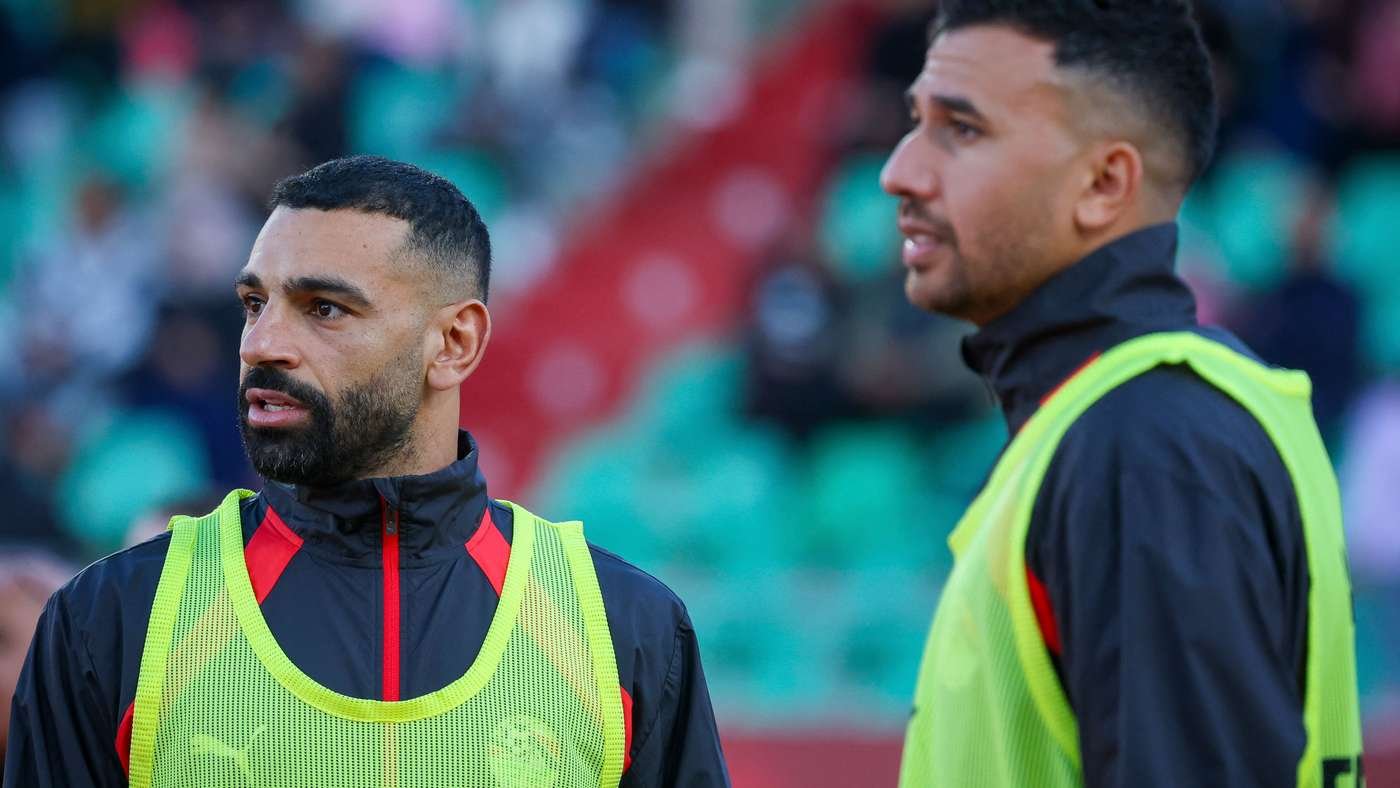 Two men in neon safety vests stand on a sports field, one with a beard looking left as the other chats nearby.