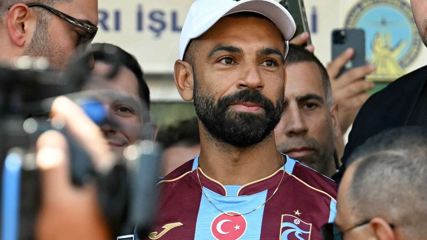 Bearded man in a white cap among a crowd taking selfies, wearing a maroon and light blue sports jersey with a crescent emblem.