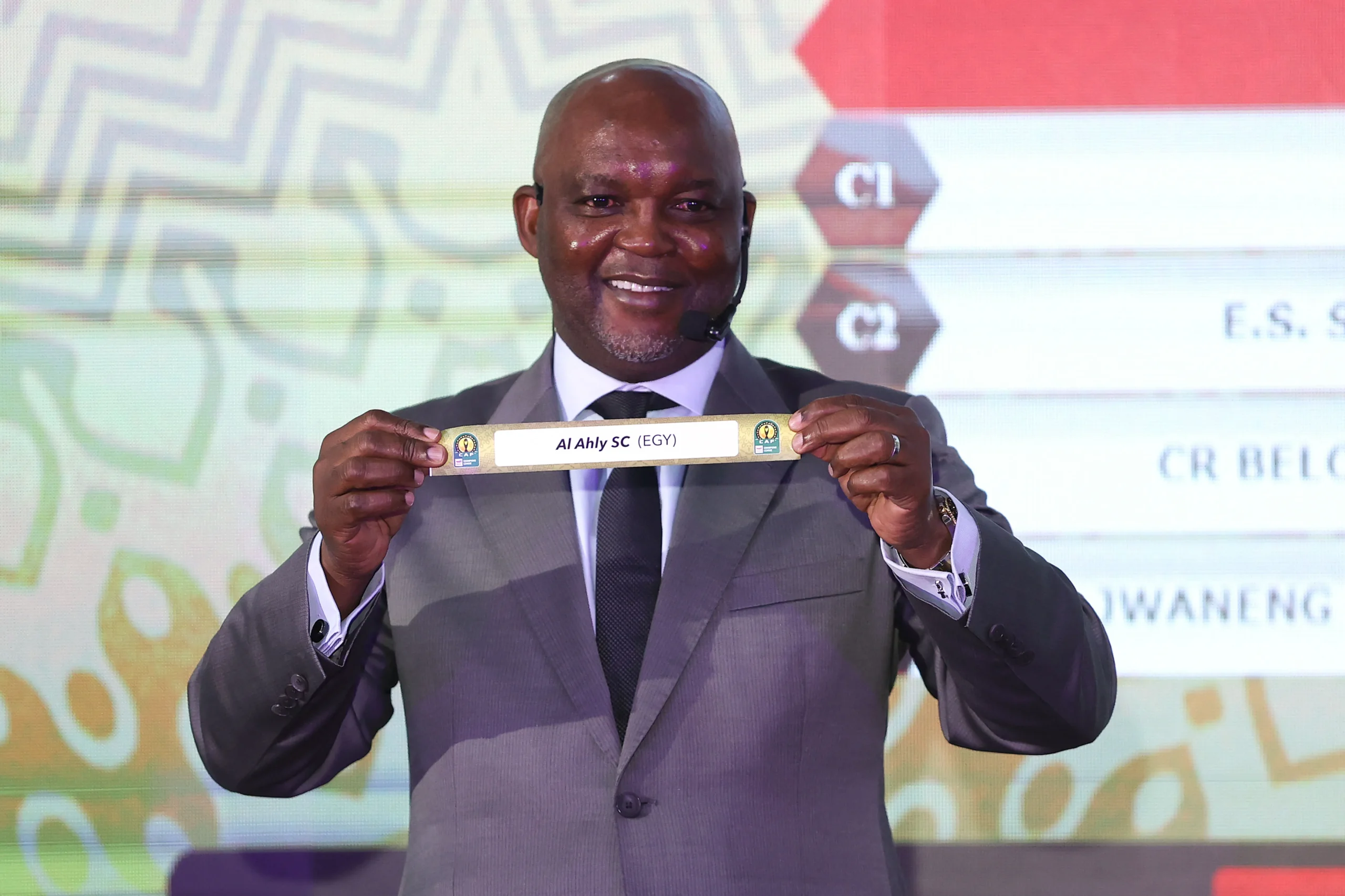 Smiling man in a gray suit holding a nameplate that reads 'Al Ahly SC (EGY)' on stage with a headset mic.