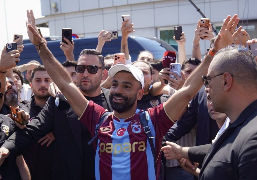 Man in a maroon and light-blue striped jersey with a white cap celebrates with arms raised as a crowd of fans takes photos around him.