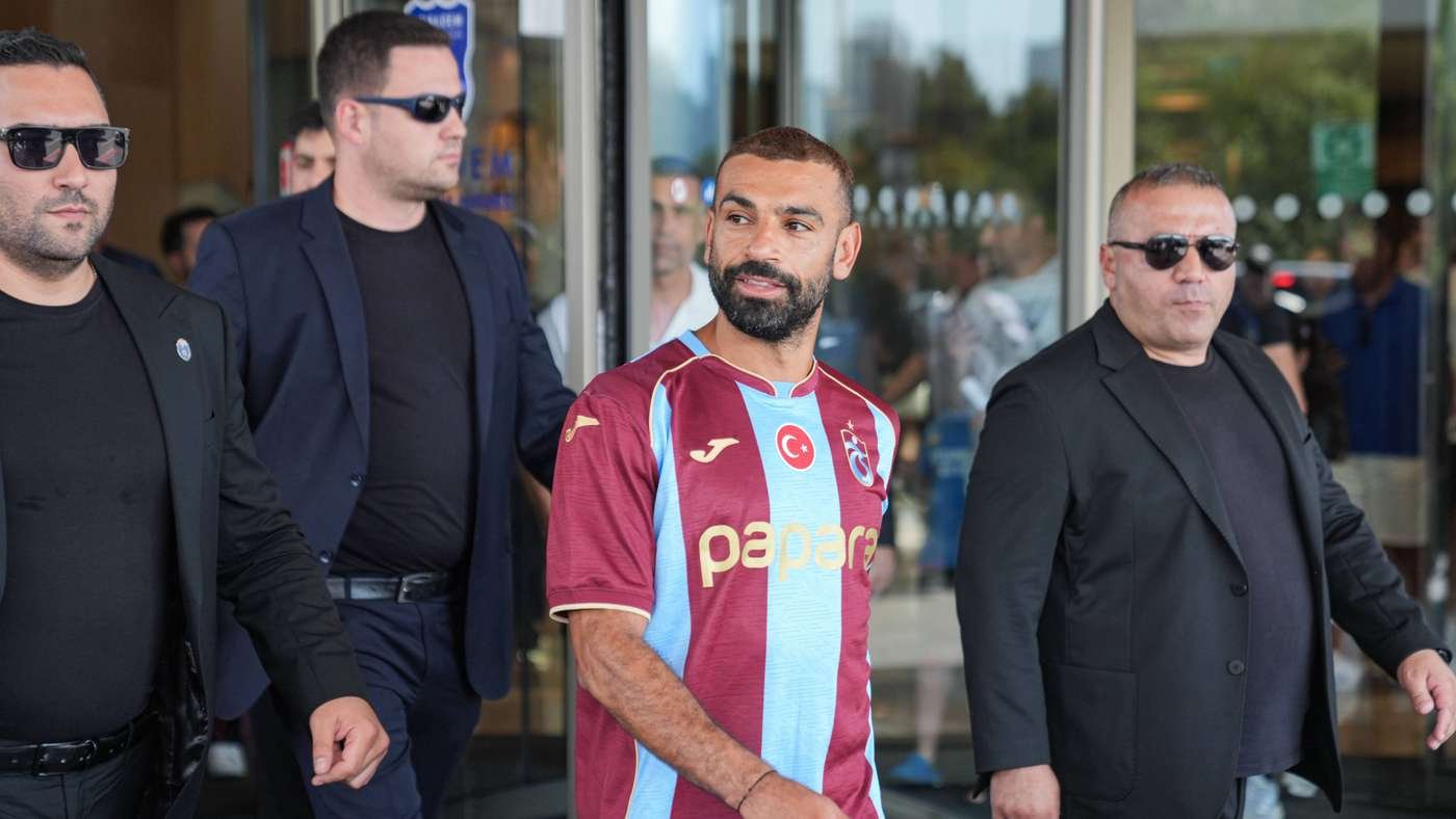 Bearded man in a maroon and light blue striped football jersey walks beside two security guards in black suits and sunglasses outside a glass entrance.