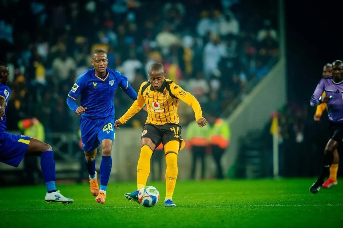 Soccer player in yellow and black shorts dribbles the ball as a blue-jersey defender closes in on a night-time match.