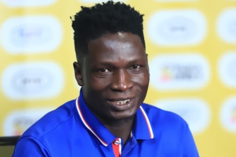 Smiling man in a blue polo shirt speaks at a press conference, with a yellow sponsor backdrop behind him.