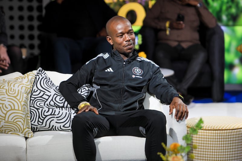 Man in a black Adidas tracksuit sits on a white couch on a stage, arms resting casually, talking to someone off-camera.