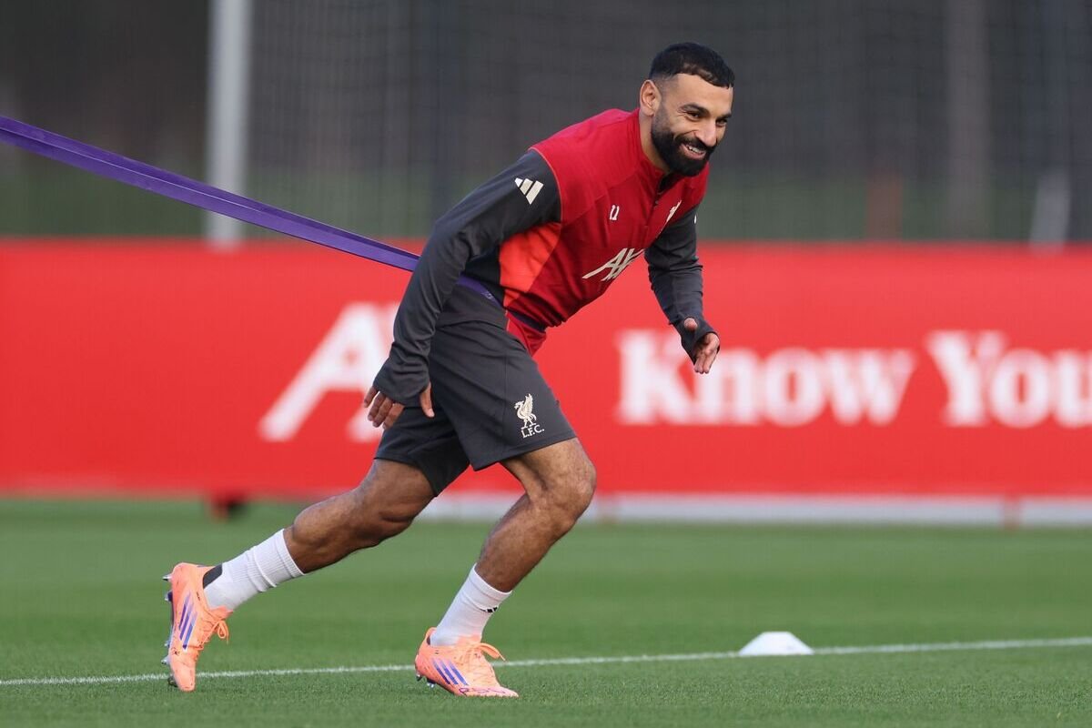 Soccer player in red training top and dark shorts sprinting on a grass field with a purple resistance band around his waist.