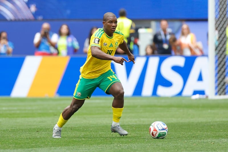 Soccer player in a yellow Brazil jersey (number 14) dribbles the ball on a green pitch with spectators and blue advertising boards in the background.