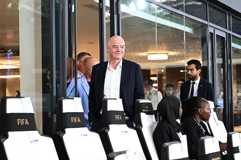Bald man in a dark suit stands beside white chairs labeled FIFA at an indoor event, with attendees seated and others in the background.