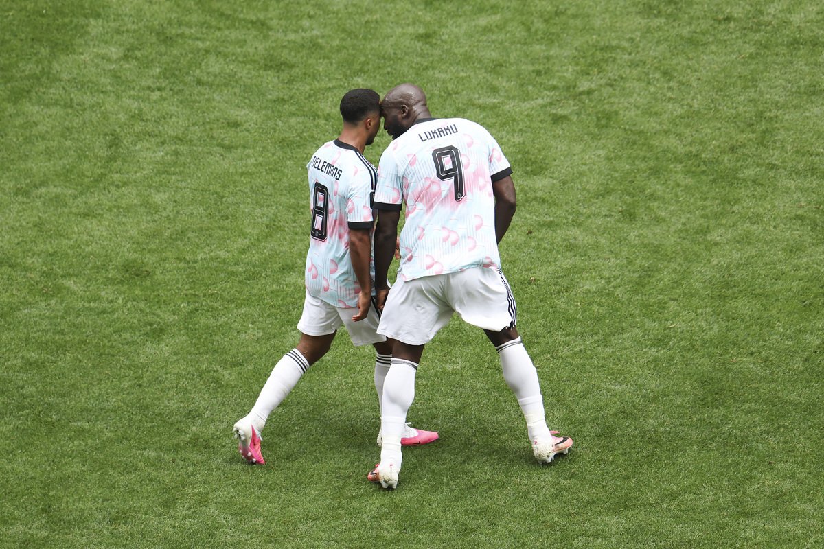 Two soccer players in white and pink kits walk together on a green grass field, with numbers 8 and 9 on their backs on the jerseys.