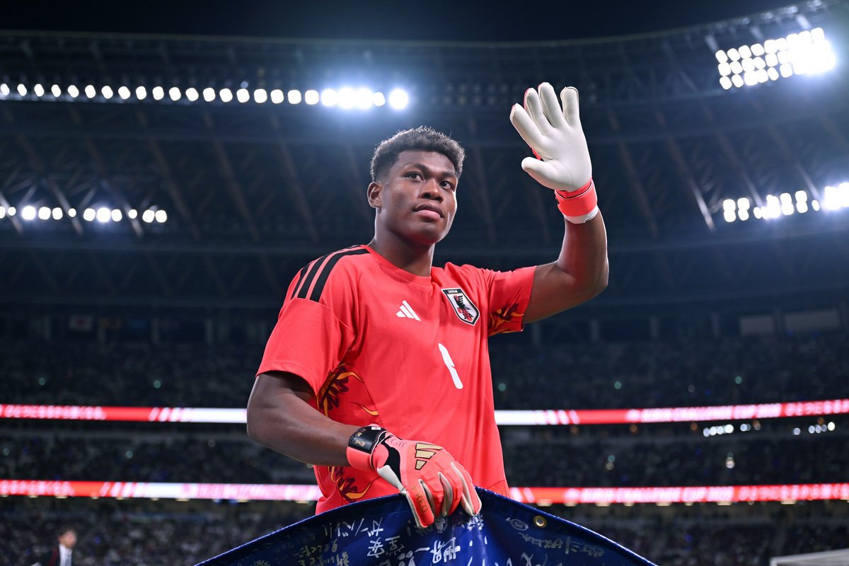 Soccer goalkeeper in red jersey and gloves raises his hand on a stadium field with bright lights in the background.