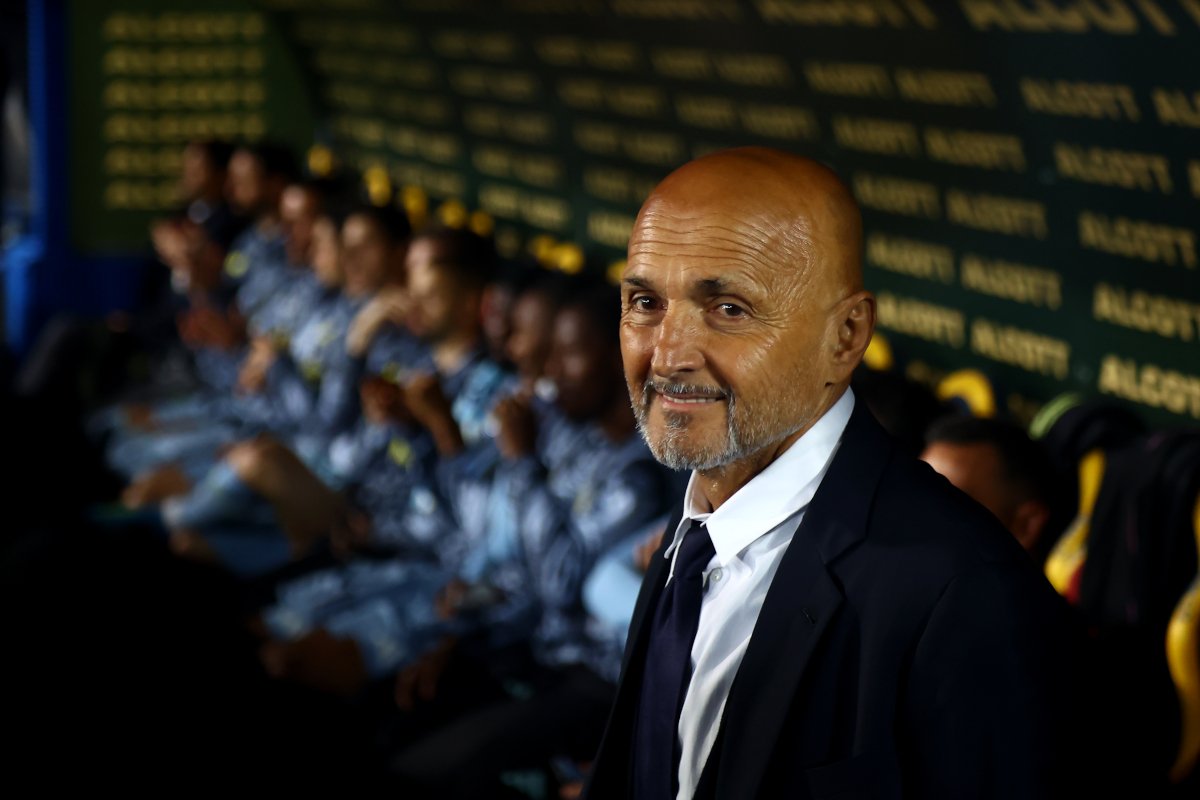 Smiling bald man in a suit seated in a dugout, with a row of players in blue uniforms behind him.