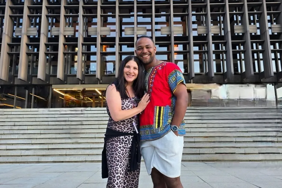A smiling couple stands arm in arm on steps in front of a modern wooden facade building, posing for a photo in bright clothing.