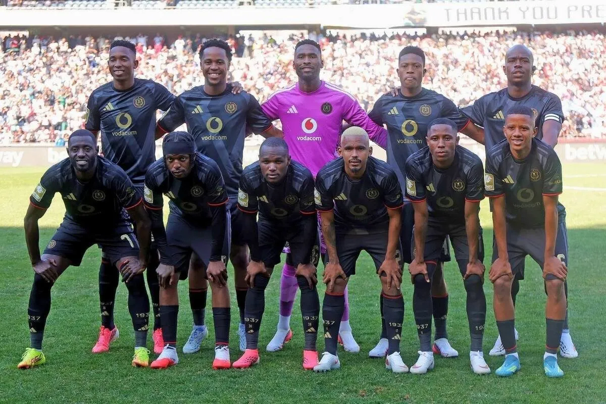 Eleven male soccer players in black uniforms pose for a team photo on a grass field with stands full of fans in the background, one in a purple goalkeeper jersey.