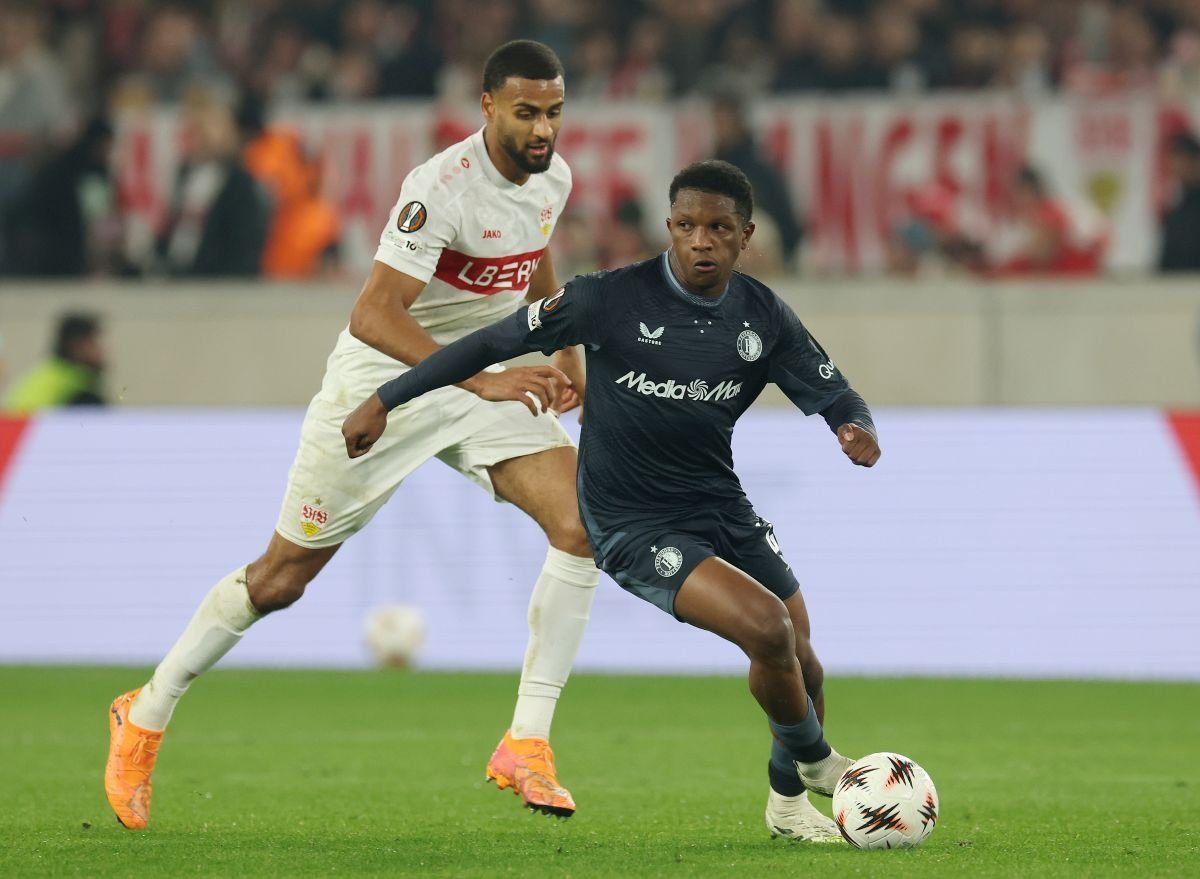 Two soccer players chase the ball during a match, one in white and one in dark blue on a green field.