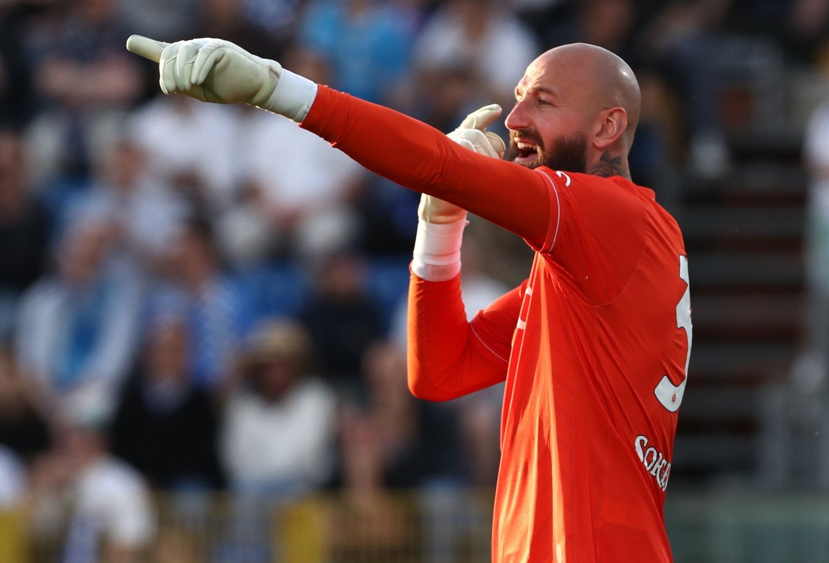 Soccer goalkeeper in a red kit points with his gloved hands as he shouts, crowd blurred in the stands behind him.